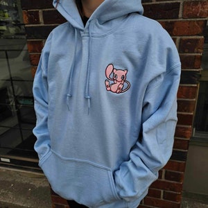 Könnte beinhalten: Ein hellblauer Hoodie mit einem gestickten rosa Comic-Charakter auf der Vorderseite. Der Charakter hat einen rosa Körper, weiße Augen und einen rosa Schwanz.