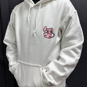 Könnte beinhalten: Weißer Kapuzenpullover mit einem gestickten rosa Comic-Charakter auf der Vorderseite.