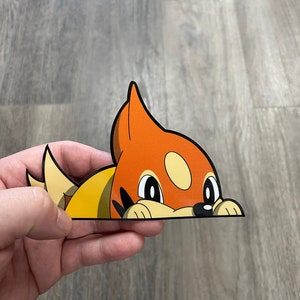May include: A die-cut sticker featuring a cartoon fox-like creature with orange, yellow, and white coloring. The sticker is peeking over a surface, with its eyes and nose visible. The sticker is held by a hand.