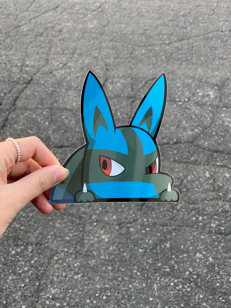 Op de afbeelding: Een sticker met een stripfiguur met blauwe oren, een blauwe streep en rode ogen. Het personage heeft een donkergroen lichaam en kijkt over een rand. De sticker wordt door een hand vastgehouden.