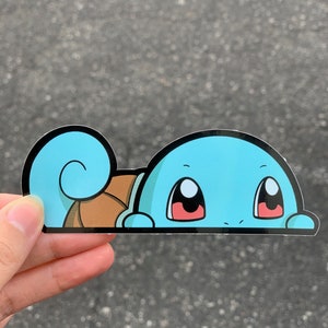 Puede incluir: Una pegatina de dibujos animados azul y marrón de un Pokémon Squirtle asomándose desde detrás de una superficie. La pegatina tiene un contorno negro y el Squirtle tiene ojos grandes y rojos.