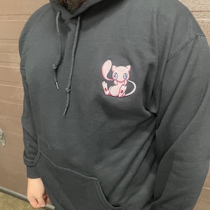 Könnte beinhalten: Schwarzer Kapuzenpullover mit einem gestickten pinkfarbenen und weißen Comic-Charakter auf der Vorderseite. Der Charakter hat einen pinkfarbenen Körper, weiße Augen und einen pinkfarbenen Schwanz.