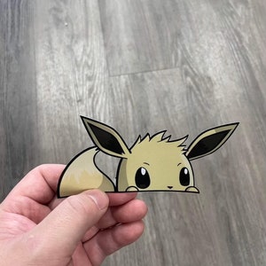 May include: A tan and black Eevee sticker, depicting the character's face and tail. The sticker is held by a hand, with the character's face peeking out from the bottom. The sticker is on a wooden surface.