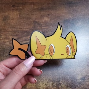 Puede incluir: Una pegatina amarilla con un personaje de dibujos animados con una marca en forma de estrella y un accesorio en forma de estrella. La pegatina tiene contornos negros y se sostiene sobre un fondo de madera.