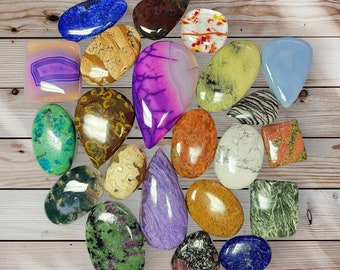 Peut inclure: Assortiment de 22 cabochons de pierres précieuses polies de différentes couleurs et motifs, notamment vert, bleu, violet, marron, rouge et blanc. Les pierres sont présentées sur une surface en bois.