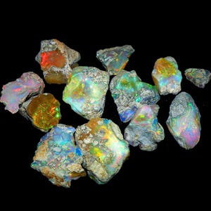 May include: A collection of rough opal gemstones, showcasing a variety of colors including red, orange, green, blue, and purple. The opals have a rough, textured surface and are set against a black background.
