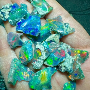 Könnte beinhalten: Eine Handvoll roher Opalsteine mit leuchtenden Farben. Die Opale weisen blaue, grüne und rote Farbblitze auf und haben eine raue, strukturierte Oberfläche. Die Steine werden in einer Hand gehalten, um ihre natürliche Schönheit zu zeigen.