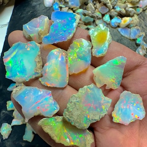 Op de afbeelding: Een verzameling ruwe opaal edelstenen, met een spectrum aan kleuren waaronder blauw, groen en flitsen van rood en geel. De opalen worden in een hand gehouden, met een achtergrond van meer opaalfragmenten.