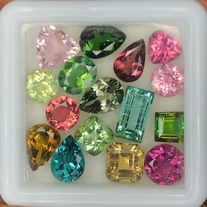 Puede incluir: Una colección de quince piedras preciosas facetadas en una caja de presentación blanca, con diversas formas como pera, redonda, cuadrada y rectangular en colores vibrantes como rosa, verde, amarillo, turquesa y rojo oscuro, sobre una base blanca.