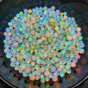 Puede incluir: Una colección de pequeñas ópalos redondos en un cuenco oscuro. Los ópalos muestran una gama de colores, incluyendo blanco, rosa, azul, verde y amarillo, con destellos iridiscentes. Los ópalos son lisos y pulidos, reflejando la luz.