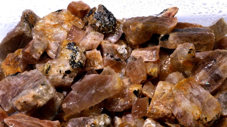 K&ouml;nnte beinhalten: Nahaufnahme eines Haufens rohen Sunstone, der die nat&uuml;rliche, rohe Sch&ouml;nheit des Edelsteins zeigt. Der Sunstone ist eine Mischung aus orange, rosa und braunen Farbt&ouml;nen, wobei einige St&uuml;cke einen leichten Schimmer haben.