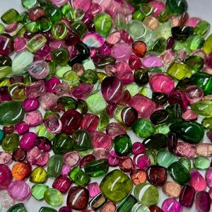 Pode incluir: Uma coleção de pedras preciosas de turmalina polidas em várias formas e tamanhos. As gemas exibem uma variedade de cores, incluindo rosa, verde e vermelho. As pedras estão espalhadas, criando uma exibição colorida e texturizada.