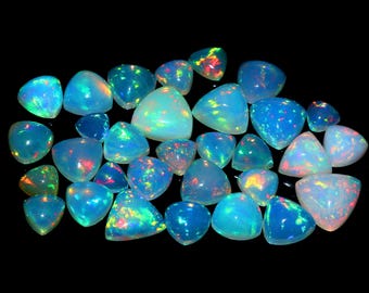 Puede incluir: Una colección de piedras preciosas de ópalo pulidas en varias formas, incluyendo triángulos y formas redondeadas. Los ópalos muestran un vibrante juego de colores con destellos de azul, verde, rojo y amarillo sobre una base blanca lechosa o translúcida, sobre un fondo negro.