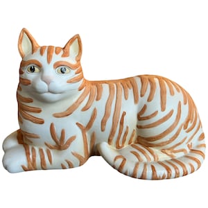 May include: A ceramic figurine of a white cat with orange stripes lying down. The cat has a realistic face with large eyes and a closed mouth.