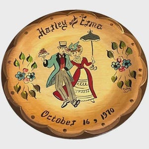 Könnte beinhalten: Ein dekorativer Vintage-Holzteller mit gewelltem Rand. Der Teller zeigt eine handgemalte Illustration eines Paares in formeller Kleidung, das einen Regenschirm und einen Kuchen hält. Der Text "Hatley and Erma" und "October 16, 1970" sind ebenfalls auf den Teller gemalt.