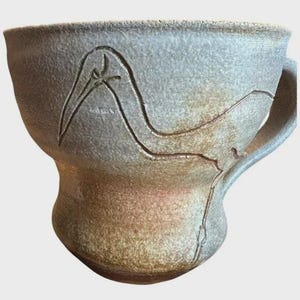 May include: A handmade ceramic mug with a bird design carved into the exterior. The mug has a wide, rounded base and a handle. The color is a mix of gray and brown, with a textured surface. The bird design is a simple, elegant outline.