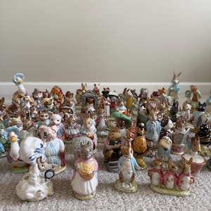 May include: A collection of ceramic figurines featuring various animal characters, including rabbits, pigs, and ducks. The figurines are painted in bright colors and are arranged on a carpeted surface. The collection includes characters in various poses and outfits.