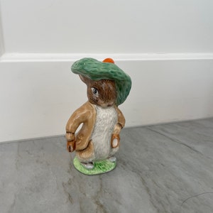 May include: A ceramic figurine of Peter Rabbit wearing a green hat with an orange flower. The figurine is standing on a green base.
