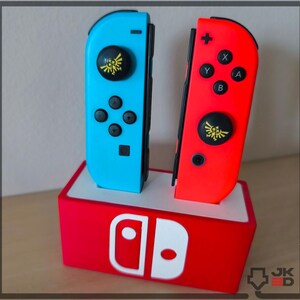 Può includere: Due controller Joy-Con per Nintendo Switch, uno blu e uno rosso, con pulsanti neri e un'emblema dorato. Sono posizionati su una base rossa e bianca con il logo Nintendo Switch.