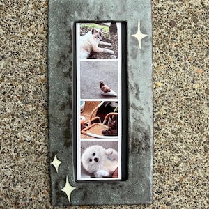 May include: A rectangular, dark gray, textured frame featuring three light-colored, four-pointed star accents. The frame holds a vertical strip of four photographs displaying a cat, a pigeon, a cat on a wicker chair, and a fluffy white dog.