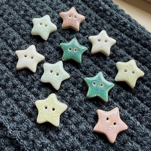Puede incluir: Una colección de diez botones con forma de estrella en varios colores, incluyendo rosa pálido, crema, verde claro y verde oscuro. Cada botón presenta un acabado de esmalte brillante y agrietado con dos orificios centrales sobre una tela gris oscuro.
