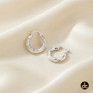 May include: A pair of silver hoop earrings featuring a twisted, rope-like texture. The earrings have a polished, reflective finish and a hinged post closure mechanism. The design consists of a continuous circular shape with a subtle spiral pattern.