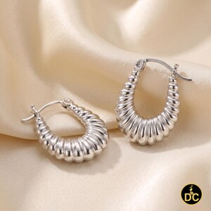 May include: A pair of silver-toned hoop earrings featuring a ribbed, croissant-like texture. The earrings have a U-shaped design with a hinged post closure. The metallic surface is polished and reflective, showing distinct vertical ridges.