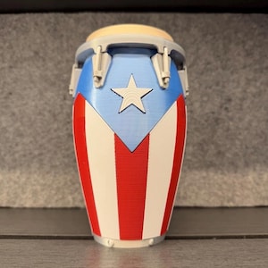 Puede incluir: Un pequeño conga decorativo con el diseño de la bandera de Puerto Rico. El tambor tiene una parte superior azul claro y una estrella blanca sobre un fondo azul. Rayas rojas y blancas adornan la parte inferior del tambor, creando una estética patriótica.