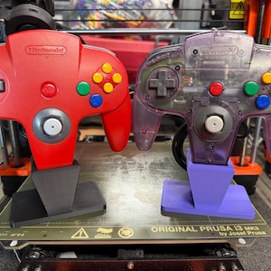 Könnte beinhalten: Zwei Nintendo 64-Controller, einer rot und einer durchscheinend lila, werden gezeigt. Der rote Controller hat ein graues Steuerkreuz und bunte Knöpfe. Der lila Controller zeigt interne Komponenten. Beide sind auf Ständern montiert.