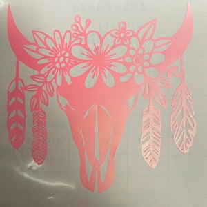 May include: Pink silhouette of a cow skull with floral accents and feathers. The skull is decorated with flowers and leaves around the top and feathers hanging from the bottom.