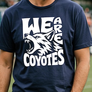Könnte beinhalten: Marineblaues T-Shirt mit weißer Grafik eines heulenden Wolfes und dem Schriftzug "WE ARE COYOTES". Das Shirt besteht aus weichem Material und eignet sich für Freizeitkleidung. Das Bild ist vor einem verschwommenen Hintergrund im Freien aufgenommen.