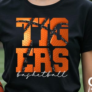 May include: Black t-shirt with the word "TIGERS" in orange, basketball-textured letters, and the word "basketball" in white script. A basketball is visible in the lower left corner.
