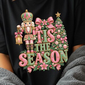 以下が含まれることがあります： ランタンを持ったくるみ割り人形と装飾されたクリスマスツリーのグラフィックが描かれた黒いTシャツ。ピンクと緑の大きなスタイリッシュな文字で「TIS THE SEASON」とプリントされており、リボンとポインセチアで装飾されています。