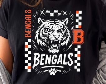 Puede incluir: Camiseta negra con un gráfico de tigre blanco y la palabra "BENGALS" en negrita. El diseño incluye detalles en naranja y una huella de pata. La letra "B" también es naranja.
