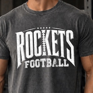 Könnte beinhalten: Ein anthrazitfarbenes Kurzarm-T-Shirt mit einem verwaschenen weißen Grafikdruck. Das Design zeigt das Wort ROCKETS, wobei ein Football den Buchstaben O ersetzt, fünf kleine Sterne darüber und das Wort FOOTBALL darunter gedruckt.