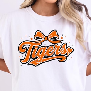 May include: A white short-sleeved t-shirt featuring a graphic design on the chest. The design displays the word Tigers in a bold orange script font with a black outline, topped with an orange bow and accented by small orange hearts and stars.