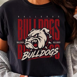 May include: A black short-sleeve t-shirt featuring a graphic print of a white bulldog head with red accents. The design includes the text BULLDOGS in large red vertical letters and white script, with the word BULLDOGS repeated in smaller text.