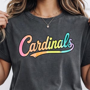Peut inclure: Un t-shirt gris anthracite à manches courtes avec le mot Cardinals imprimé dans une police cursive en dégradé multicolore passant du rose au jaune et au bleu, avec une petite icône de cœur au-dessus de la lettre i.
