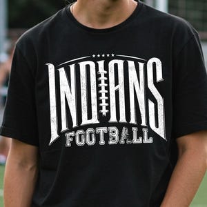 Könnte beinhalten: Ein schwarzes Kurzarm-T-Shirt mit einem weißen, abgenutzten Grafikdruck, der INDIANS zeigt, wobei ein Football-Schnürsenkel-Design den Buchstaben A ersetzt, sowie das Wort FOOTBALL darunter, gekrönt von fünf kleinen weißen Sternen.