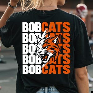 May include: A black short-sleeve t-shirt featuring a graphic on the back with the word BOBCATS repeated five times in white and orange text, overlaid with a stylized illustration of a snarling bobcat head in orange, white, and black.
