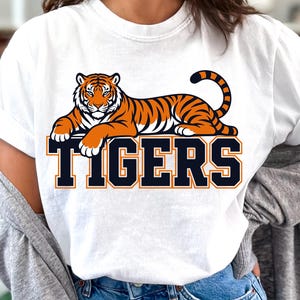 Pode incluir: Camiseta branca com um desenho de tigre laranja e preto. A palavra "TIGERS" é impressa em azul marinho com bordas laranja sob o tigre. A camiseta é de estilo casual.