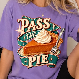 Puede incluir: Una camiseta de manga corta de color morado claro con un gráfico de una porción de tarta de calabaza con nata montada y un tenedor de plata. El texto "PASS THE PIE" aparece en una fuente retro sobre un diseño de cinta azul verdosa.