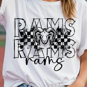 May include: White t-shirt with a black and white graphic design. The design features the word "RAMS" in a stylized font, a ram's head, and a checkered pattern. The shirt is worn by a person wearing sunglasses.