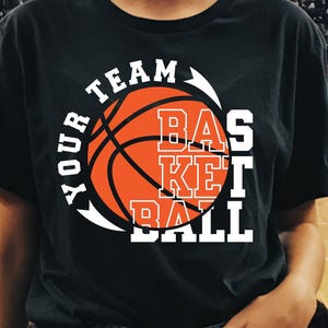 Könnte beinhalten: Schwarzes T-Shirt mit dem Text "YOUR TEAM BASKETBALL" in Weiß und Orange. Das Design zeigt eine Basketball-Grafik. Das T-Shirt wird von einer Person in einer Basketballarena getragen.