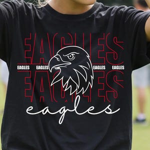 Op de afbeelding: Zwart T-shirt met een witte adelaarskop en het woord "EAGLES" in rood en wit. Het woord "EAGLES" wordt meerdere keren herhaald in een gestapeld ontwerp. Het shirt wordt gedragen met een spijkerbroek.