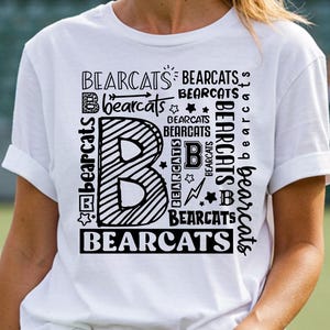 Puede incluir: Camiseta blanca con la palabra "BEARCATS" en negrita negra. La letra "B" se muestra de forma destacada con un diseño a rayas. Texto y gráficos adicionales de "BEARCATS" rodean el diseño principal.