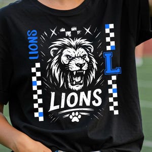 May include: Black t-shirt with a white and blue graphic of a roaring lion, the word "LIONS", and a paw print. The word "L" is in blue. The shirt has a crew neck and short sleeves. The background is a green field.