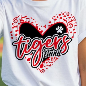 May include: White t-shirt with a heart-shaped design featuring musical notes and the words "tigers Band" in red and black. A paw print is visible within the heart. The shirt is made of a soft material.