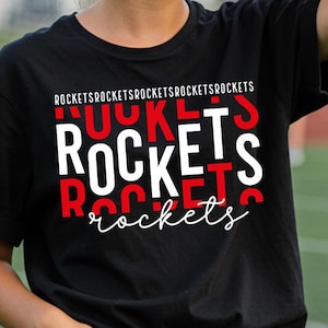 May include: Black t-shirt with the word "ROCKETS" in white and red block letters. The word "rockets" is repeated in a script font. The shirt is worn by a person in blue jeans.