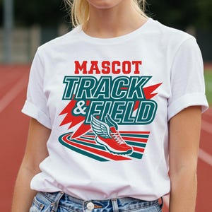 May include: White t-shirt with the text "MASCOT TRACK & FIELD" in red and teal. The shirt features a graphic of a running shoe with wings. The person is wearing blue denim shorts and a white watch.